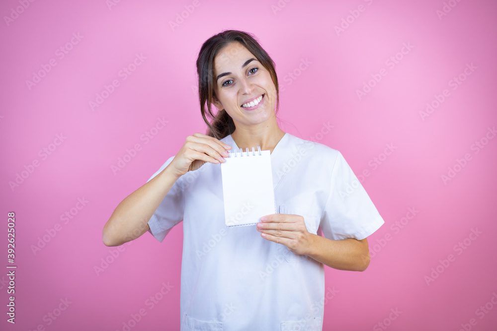 Young brunette doctor girl wearing nurse or surgeon uniform over isolated pink background smiling and showing blank notebook in her hand Young brunette doctor girl wearing nurse or surgeon uniform over isolated pink background smiling and showing blank notebook in her hand