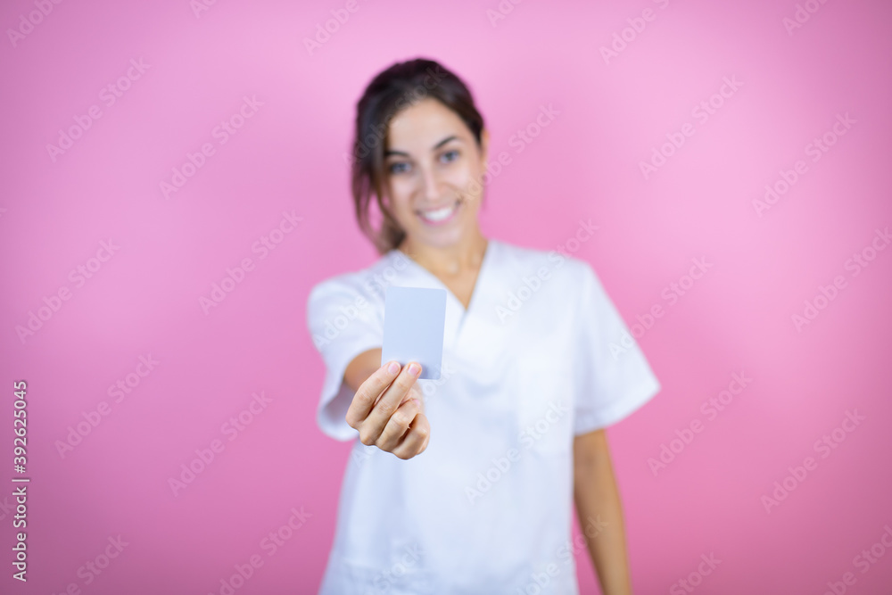 Young brunette doctor girl wearing nurse or surgeon uniform over isolated pink background smiling and holding white card Young brunette doctor girl wearing nurse or surgeon uniform over isolated pink background smiling and holding white card