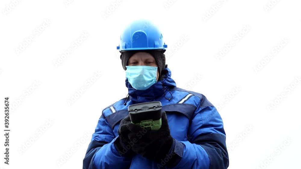 Male engineer surveyor in blue helmet and winter jacket stands in protective medical mask from coronavirus against a bleached sky and manipulates the tablet from global navigation satellite system. Male engineer surveyor in blue helmet and winter jacket stands in protective medical mask from coronavirus against a bleached sky and manipulates the tablet from global navigation satellite system.