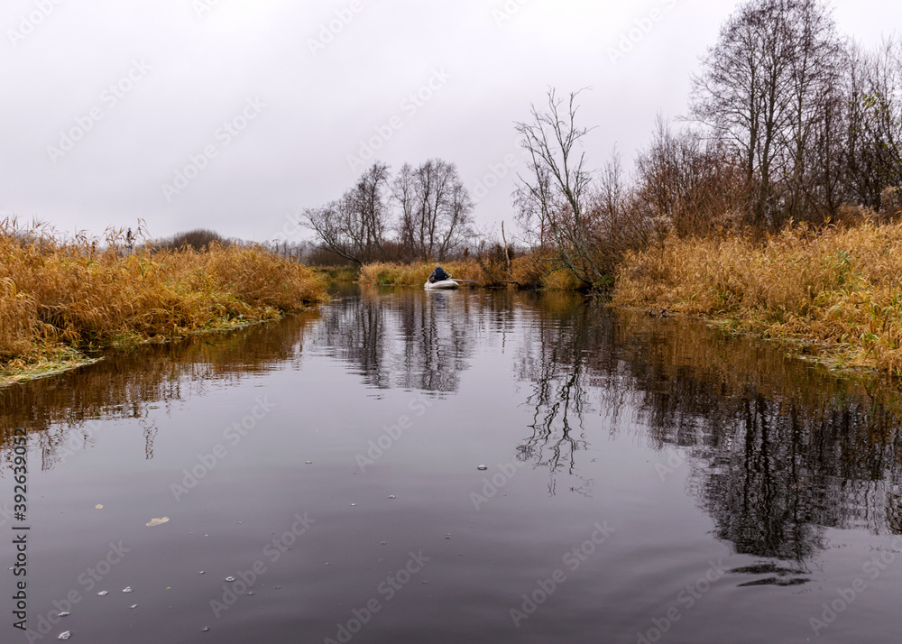 gray and cloudy day, fisherman in a white boat, river bank with bare trees and bushes, shore reflection in river water