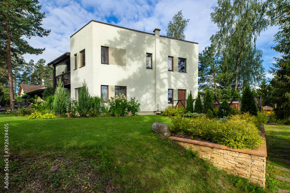 Modern exterior of luxary private house. Garden with pines, thujas and flowers. Green lawn. Cloudy blue sky. Modern exterior of luxary private house. Garden with pines, thujas and flowers. Green lawn. Cloudy blue sky.