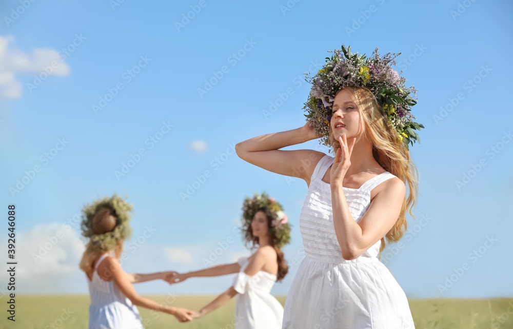 Young women wearing wreaths made of beautiful flowers in field on sunny day Young women wearing wreaths made of beautiful flowers in field on sunny day