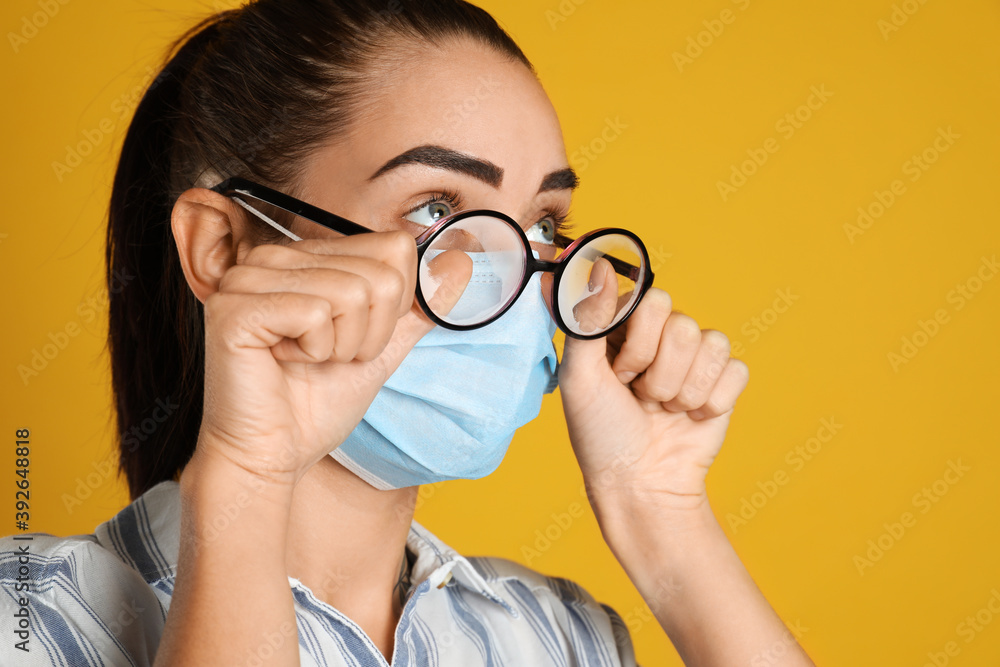 Woman wiping foggy glasses caused by wearing medical mask on yellow background, closeup Woman wiping foggy glasses caused by wearing medical mask on yellow background, closeup