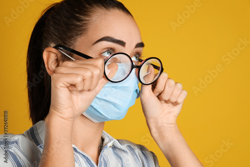 Fototapeta Naklejka Na Ścianę i Meble -  Woman wiping foggy glasses caused by wearing medical mask on yellow background, closeup