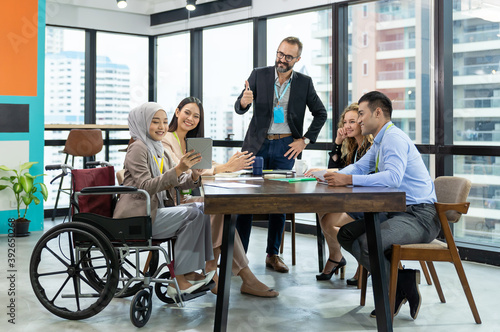 Asian Muslim business woman in hijab headscarf sitting on wheelchair presenting of her work to corporate colleagues in meeting in the modern office. diverse corporate colleagues and multicultural