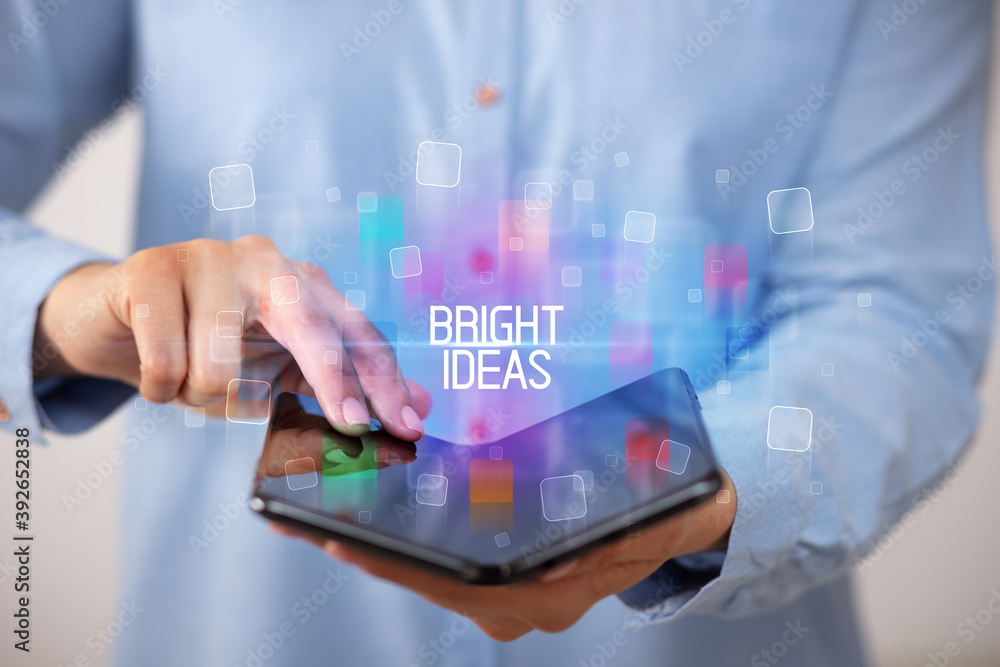 Young man holding a foldable smartphone with BRIGHT IDEAS inscription, educational concept Young man holding a foldable smartphone with BRIGHT IDEAS inscription, educational concept
