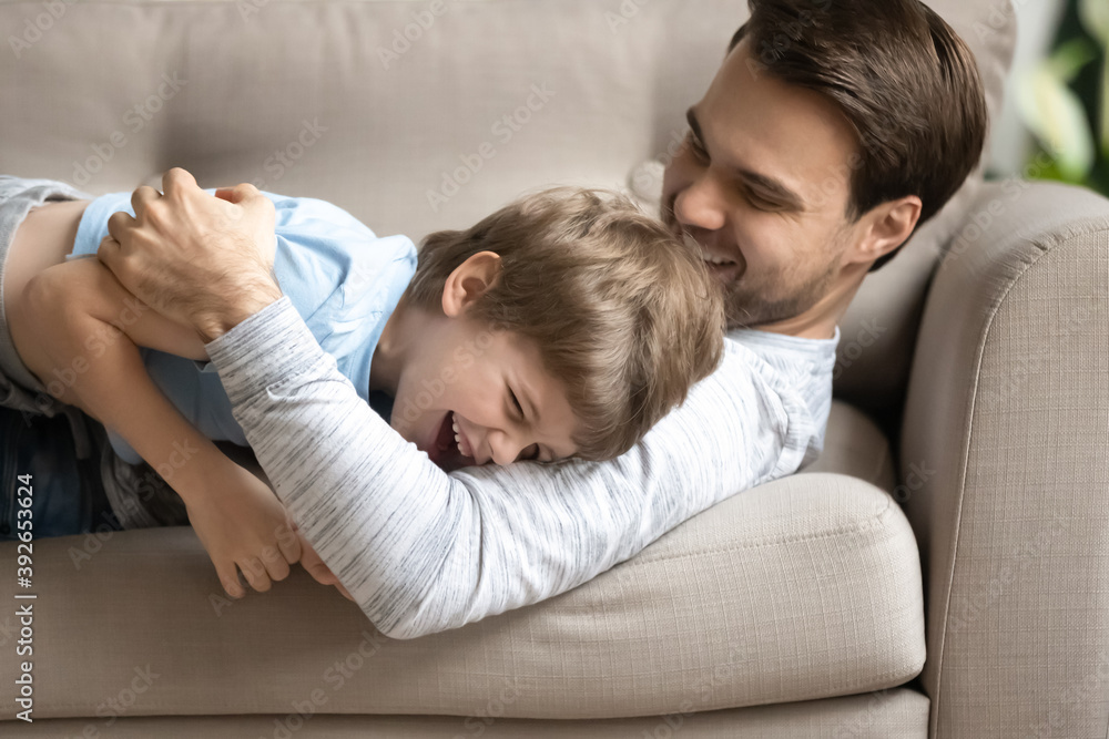 Close up overjoyed young father and adorable son hugging, lying on cozy couch at home, excited loving dad and little boy child cuddling, relaxing on sofa, family enjoying leisure time together Close up overjoyed young father and adorable son hugging, lying on cozy couch at home, excited loving dad and little boy child cuddling, relaxing on sofa, family enjoying leisure time together