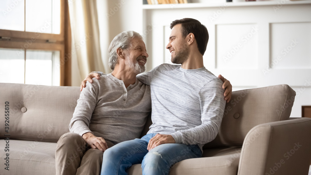 Happy mature father and adult son chatting, sitting on cozy couch in living room, smiling older grandfather with grandson enjoying pleasant conversation, leisure time at home, two generations Happy mature father and adult son chatting, sitting on cozy couch in living room, smiling older grandfather with grandson enjoying pleasant conversation, leisure time at home, two generations