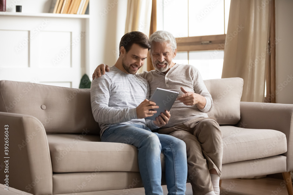 Happy mature father and adult son using tablet at home together, smiling older grandfather and grandson looking at device screen, sitting on couch, young man teaching senior dad to use computer Happy mature father and adult son using tablet at home together, smiling older grandfather and grandson looking at device screen, sitting on couch, young man teaching senior dad to use computer
