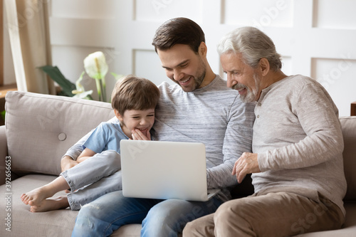 Smiling little boy with father and mature grandfather using laptop, sitting on couch, happy three generations of men looking at computer screen together, watching video, shopping or chatting online