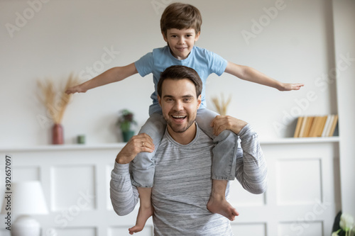 Funny portrait little boy pretending flying with hands outstretched sitting on fathers shoulders, family enjoying leisure time, playing active game, smiling dad and adorable son looking at camera