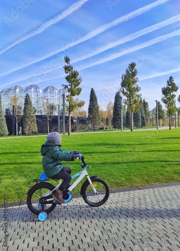 a boy on a bicycle rides up a hill along a path in a recreation park against the background of a waterfall, green grass and looks at the white footprints from the plane against the blue sky
