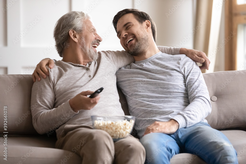 Happy older father with son hugging, laughing, watching tv at home, enjoying leisure time together, overjoyed mature grandfather with grandson cuddling, sitting on couch with popcorn snack Happy older father with son hugging, laughing, watching tv at home, enjoying leisure time together, overjoyed mature grandfather with grandson cuddling, sitting on couch with popcorn snack