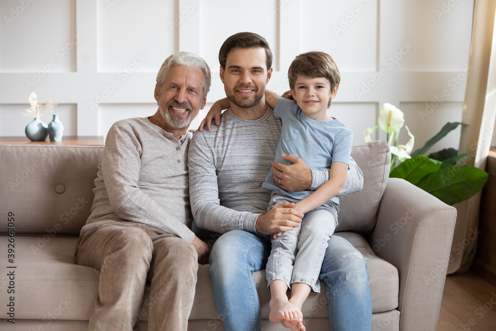 Family portrait happy three generations of men sitting on couch, hugging, smiling young father holding adorable son, little boy with dad and grandfather looking at camera, posing for photo at home Family portrait happy three generations of men sitting on couch, hugging, smiling young father holding adorable son, little boy with dad and grandfather looking at camera, posing for photo at home