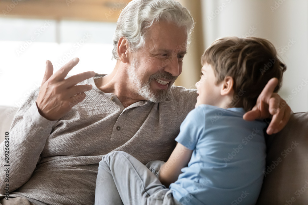 Close up loving mature grandfather chatting with adorable grandson, having pleasant conversation, happy older man and little boy grandchild enjoying leisure time together, sitting on couch at home