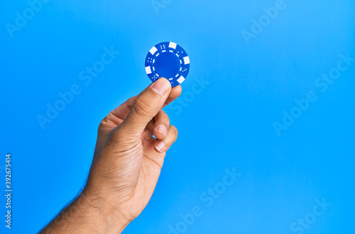 Hand of hispanic man holding casino chip over isolated blue background.