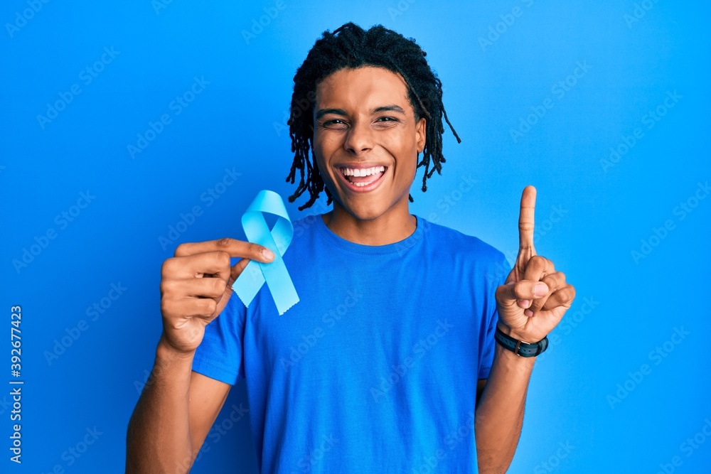 Young african american man holding blue ribbon smiling with an idea or question pointing finger with happy face, number one Young african american man holding blue ribbon smiling with an idea or question pointing finger with happy face, number one