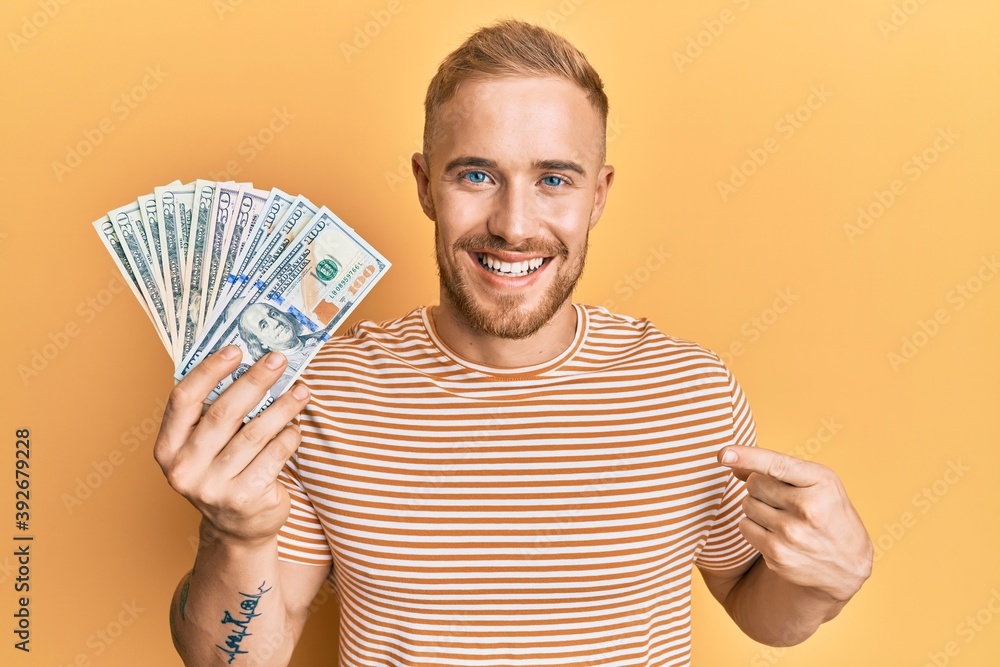 Young caucasian man holding dollars smiling happy pointing with hand and finger Young caucasian man holding dollars smiling happy pointing with hand and finger