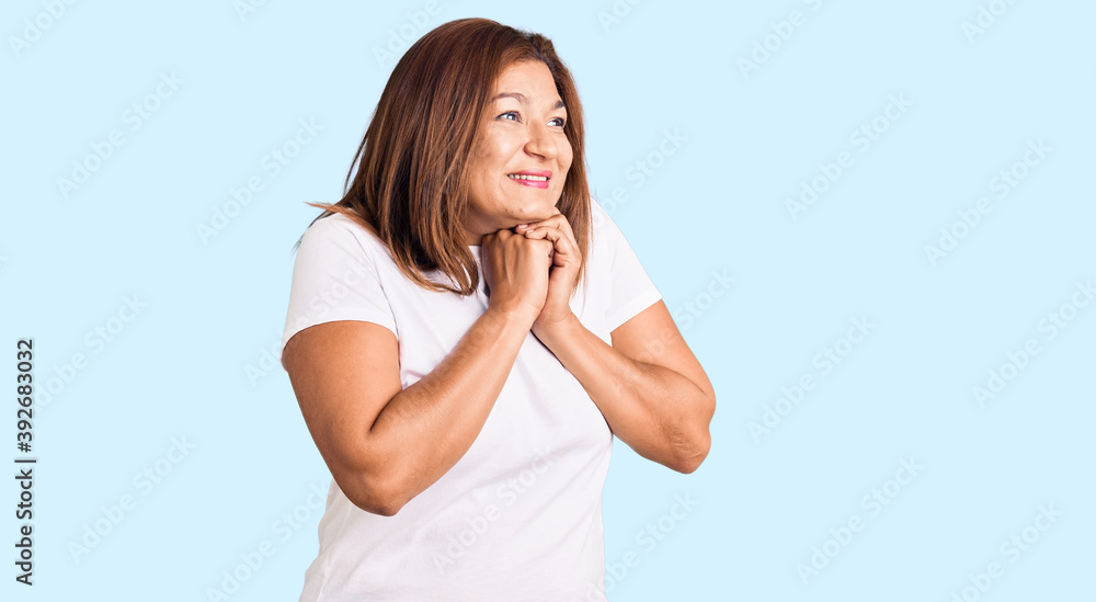 Middle age latin woman wearing casual white tshirt laughing nervous and excited with hands on chin looking to the side