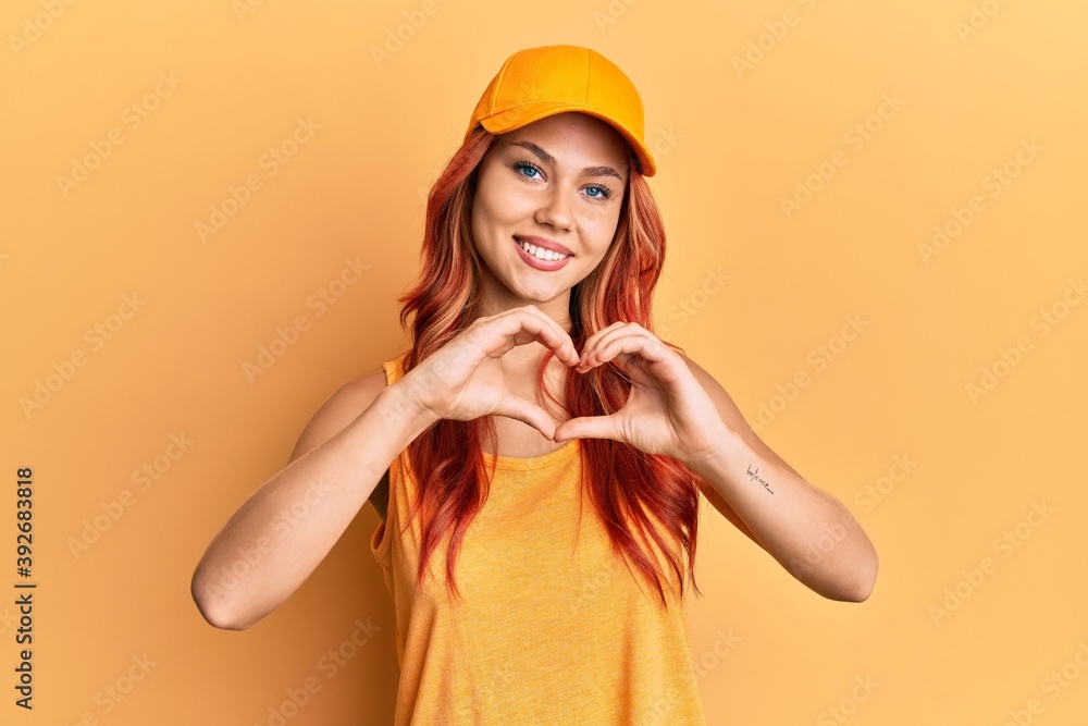 Young beautiful redhead woman wearing casual clothes and yellow cap smiling in love doing heart symbol shape with hands. romantic concept. Young beautiful redhead woman wearing casual clothes and yellow cap smiling in love doing heart symbol shape with hands. romantic concept.