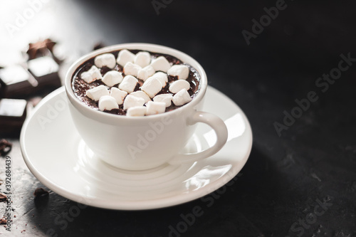 Hot chocolate drink in white cup with marshmallow, broken chocolate cubes and star anise on dark background