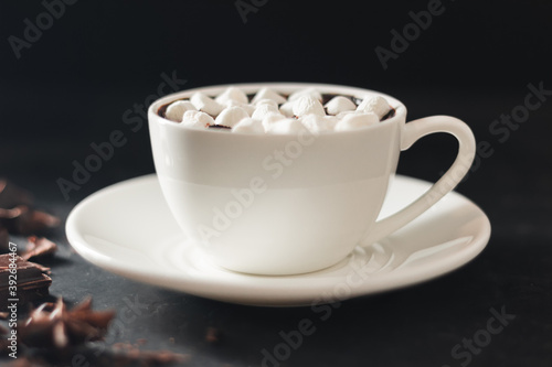 Hot chocolate drink in white cup with marshmallow  broken chocolate cubes and star anise on dark background