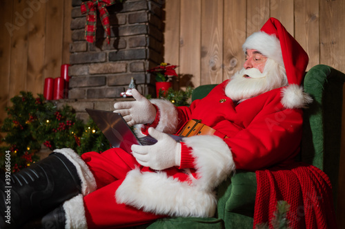 Happy santa claus sits at home in an armchair and remotely wishes merry christmas via video chat on a laptop