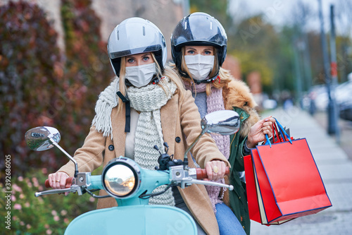 Two women wearing masks and holding shopping bags on scooter
