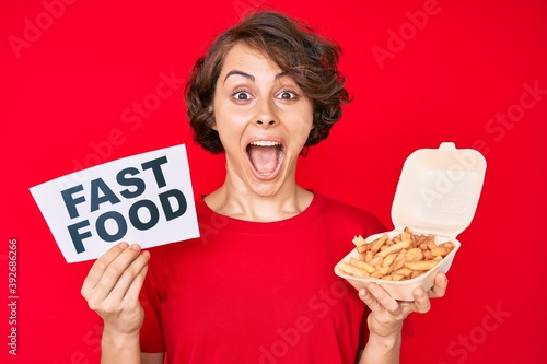 Young hispanic woman holding potato chip and fast food banner celebrating crazy and amazed for success with open eyes screaming excited.