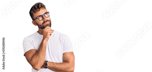 Young hispanic man wearing casual clothes and glasses with hand on chin thinking about question, pensive expression. smiling with thoughtful face. doubt concept. © Krakenimages.com