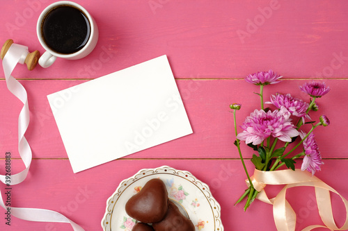 Woman table top view. Blank greeting card, envelope, coffee cup, ribbon, flowers, heart-shaped chocolate on pastel pink background. Female desk with feminine accessories. Top view, flat lay