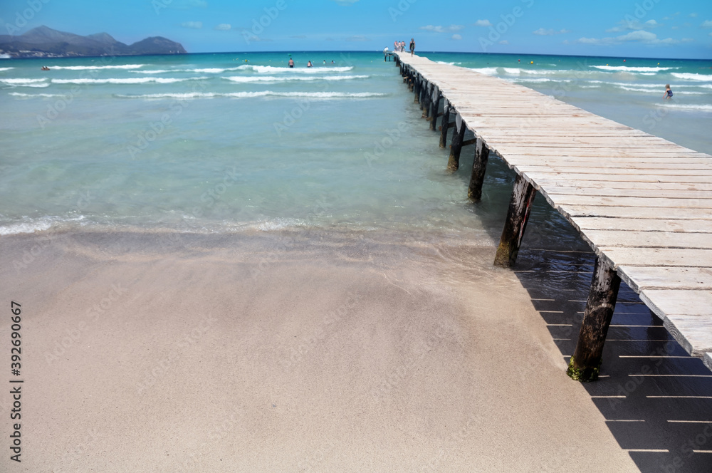 Summer day by the Alcudia beach, Mallorca, Spain. Tourists swimming and walking on the wooden bridge.