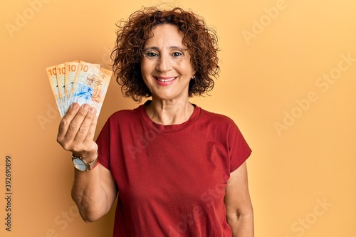 Beautiful middle age mature woman holding 10 swiss franc banknotes looking positive and happy standing and smiling with a confident smile showing teeth