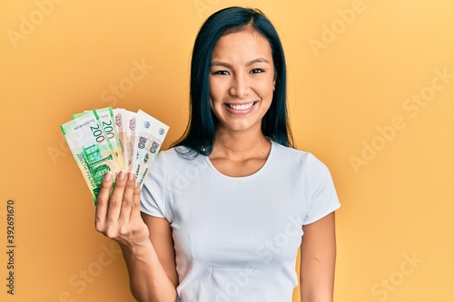 Beautiful hispanic woman holding russian ruble banknotes looking positive and happy standing and smiling with a confident smile showing teeth