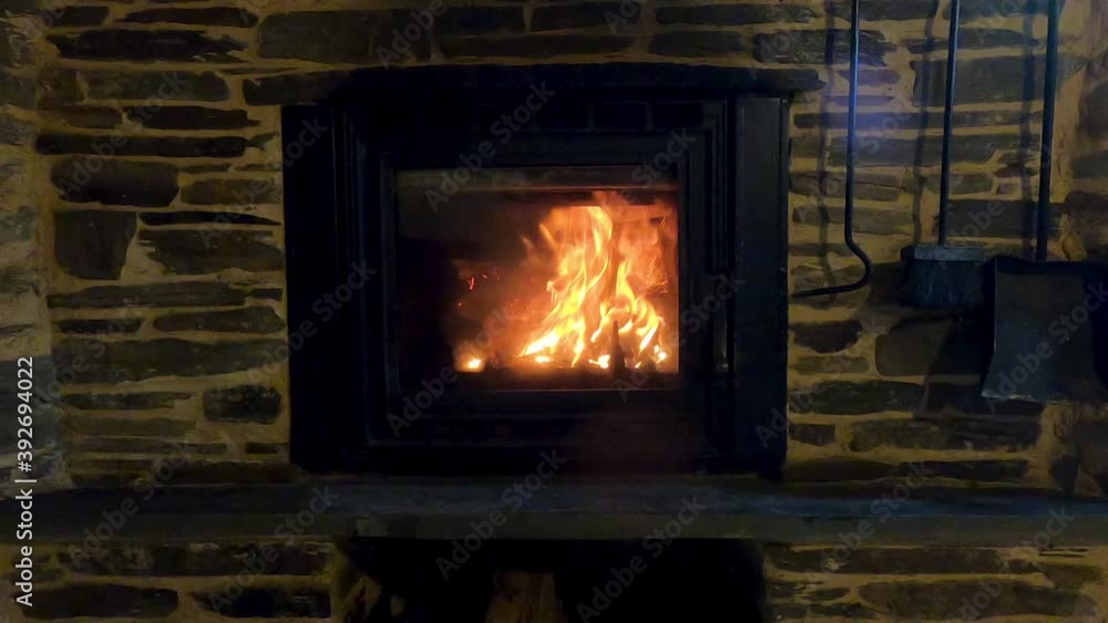 Beautiful rustic iron fireplace, surrounded by a stone wall, lighting and warming the room with its large and orange flames, 4k Beautiful rustic iron fireplace, surrounded by a stone wall, lighting and warming the room with its large and orange flames, 4k