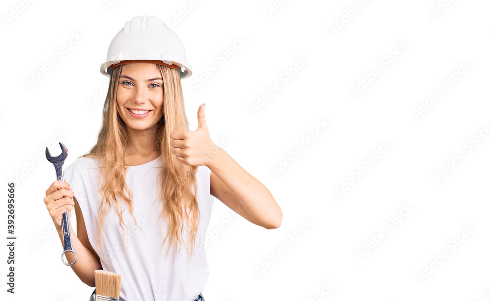 Beautiful caucasian woman with blonde hair wearing hardhat and builder clothes smiling happy and positive, thumb up doing excellent and approval sign