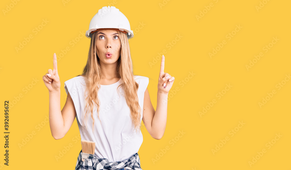 Beautiful caucasian woman with blonde hair wearing hardhat and painter clothes amazed and surprised looking up and pointing with fingers and raised arms. Beautiful caucasian woman with blonde hair wearing hardhat and painter clothes amazed and surprised looking up and pointing with fingers and raised arms.