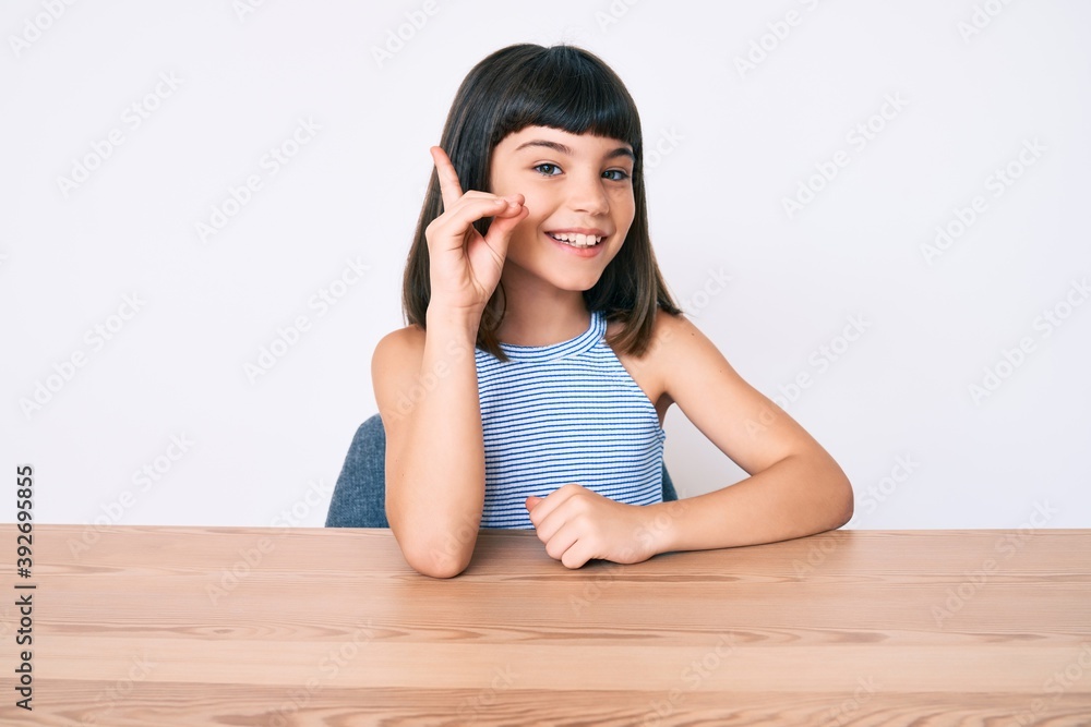 Young little girl with bang wearing casual clothes sitting on the table smiling with an idea or question pointing finger up with happy face, number one Young little girl with bang wearing casual clothes sitting on the table smiling with an idea or question pointing finger up with happy face, number one