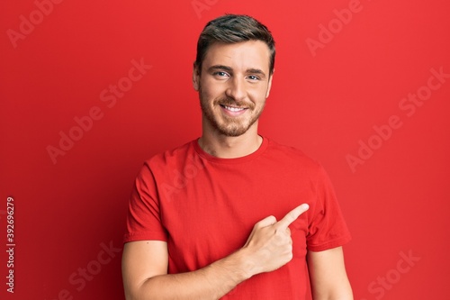 Handsome caucasian man wearing casual red tshirt cheerful with a smile on face pointing with hand and finger up to the side with happy and natural expression