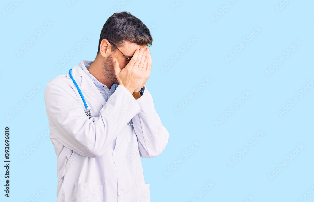 Handsome young man with bear wearing doctor uniform with sad expression covering face with hands while crying. depression concept.