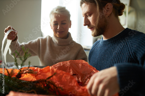 Family bonds  togetherness  holidays and celebration. Portrait of stylish bearded man sitting at home with his elderly mother wrapping Christmas presents using decorative paper and natural fir twigs