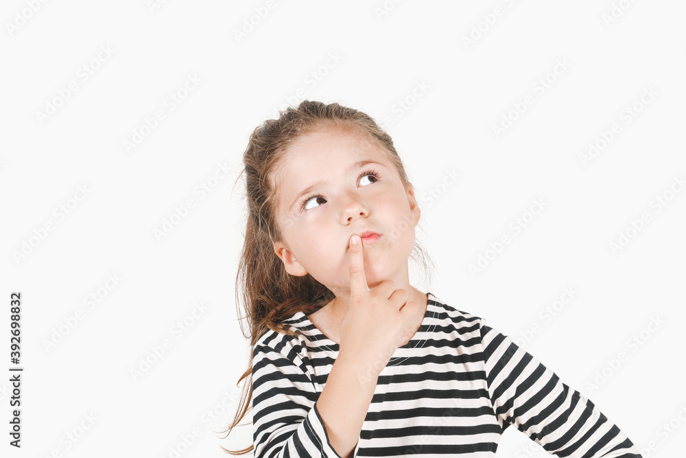 Thoughtful girl looking up aside and touching her mouth with finger. Girl with eyes directed sideways and head tilted to one side. Posing little girl wearing striped shirt. Isolated background. Thoughtful girl looking up aside and touching her mouth with finger. Girl with eyes directed sideways and head tilted to one side. Posing little girl wearing striped shirt. Isolated background.