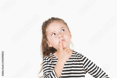 Thoughtful girl looking up aside and touching her mouth with finger. Girl with eyes directed sideways and head tilted to one side. Posing little girl wearing striped shirt. Isolated background.