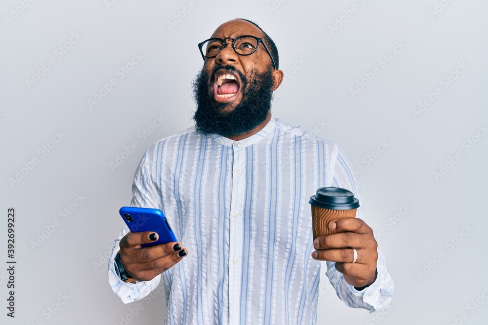 Young african american man using smartphone and drinking a cup of coffee angry and mad screaming frustrated and furious, shouting with anger looking up.