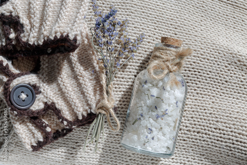 A glass bottle of sea salt with a cork for a relaxing bath on a beige textile knitted background next to a small bouquet of lavender and a body scrub