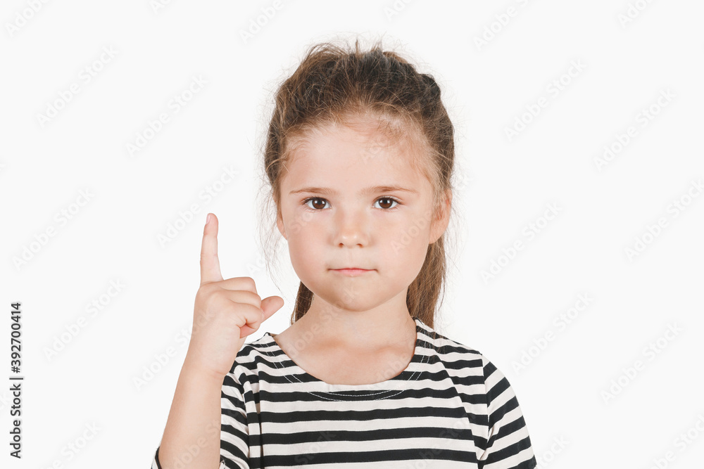 Serious girl pointing finger up, showing Number one. Close up of posing little girl wearing a striped shirt, looking at the camera on an isolated background. First, the winner, attention concept. Serious girl pointing finger up, showing Number one. Close up of posing little girl wearing a striped shirt, looking at the camera on an isolated background. First, the winner, attention concept.