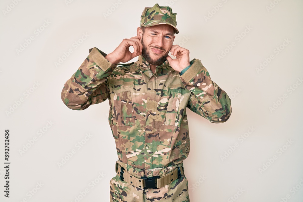 Young caucasian man wearing camouflage army uniform covering ears with fingers with annoyed expression for the noise of loud music. deaf concept. Young caucasian man wearing camouflage army uniform covering ears with fingers with annoyed expression for the noise of loud music. deaf concept.