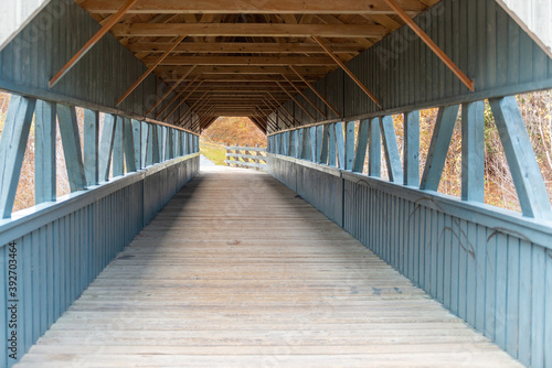 A wooden footbridge with a wood roof covering. The vintage bridge has a wood deck, blue wood sides and a natural wood ceiling. The sides are partially open. There are autumn trees in the background.