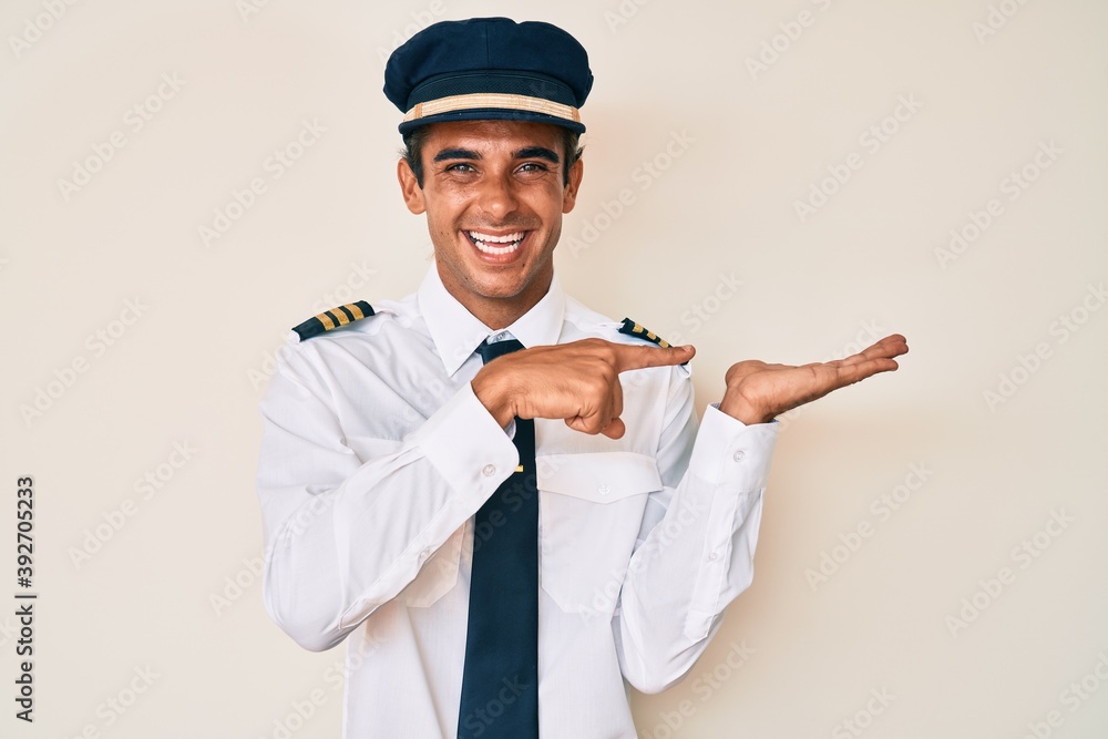 Young hispanic man wearing airplane pilot uniform amazed and smiling to the camera while presenting with hand and pointing with finger. Young hispanic man wearing airplane pilot uniform amazed and smiling to the camera while presenting with hand and pointing with finger.
