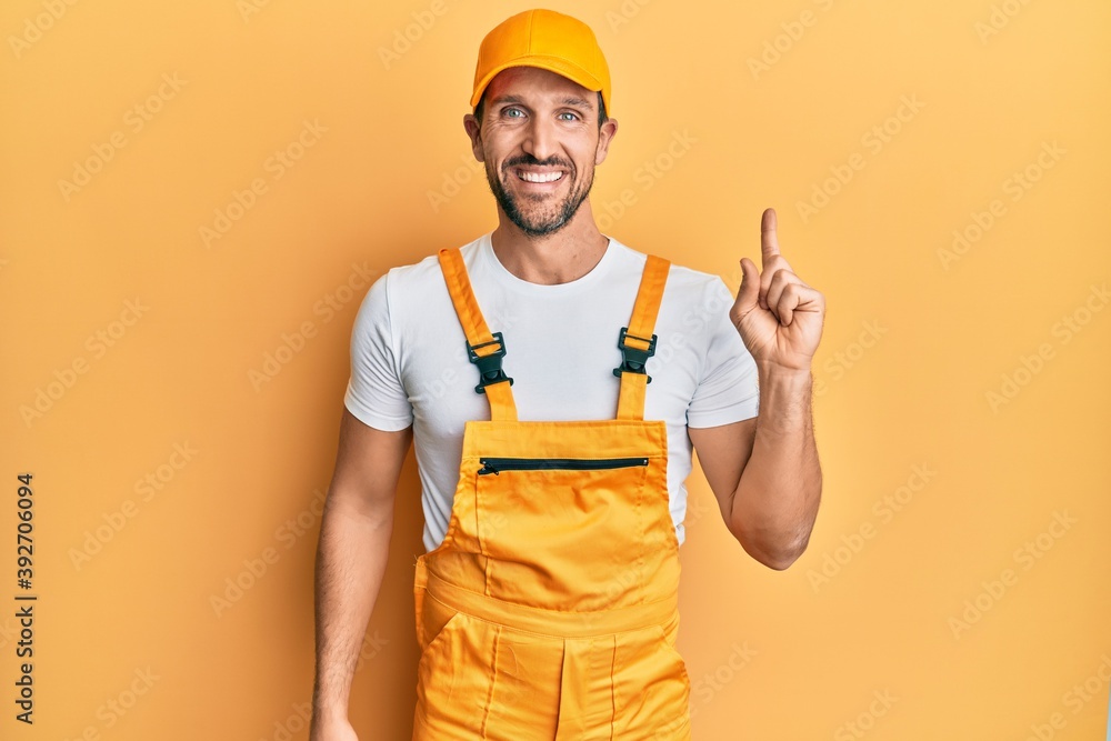Young handsome man wearing handyman uniform over yellow background with a big smile on face, pointing with hand finger to the side looking at the camera. Young handsome man wearing handyman uniform over yellow background with a big smile on face, pointing with hand finger to the side looking at the camera.